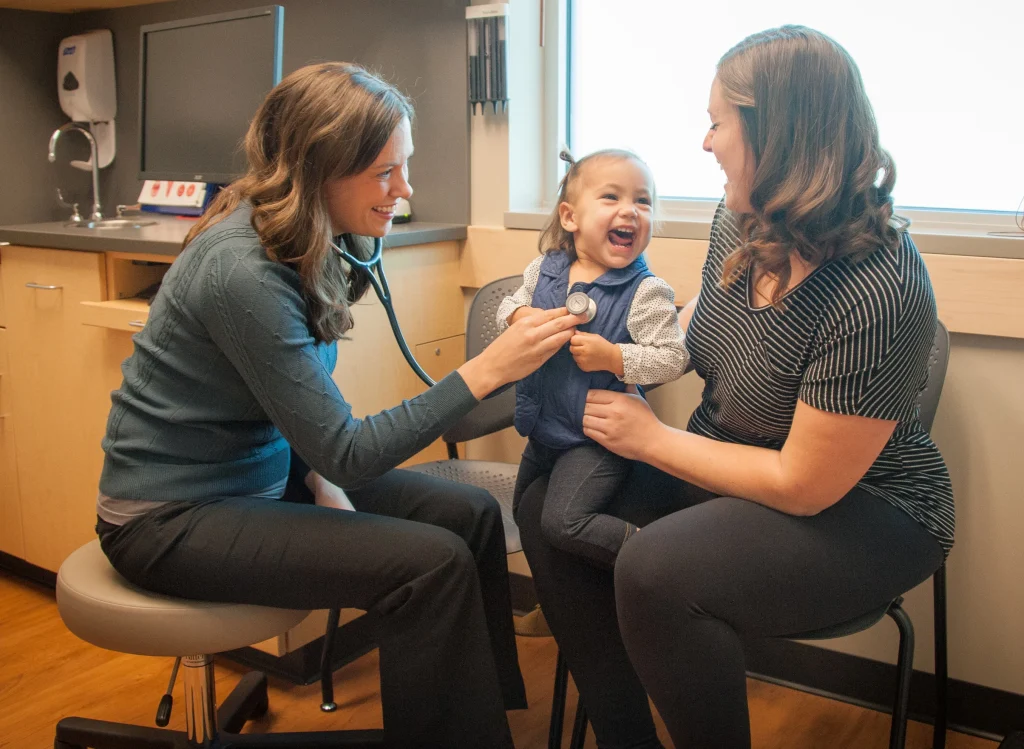 Photo of a smiling doctor with a small chid patient sitting in their mother's lap, laughing .