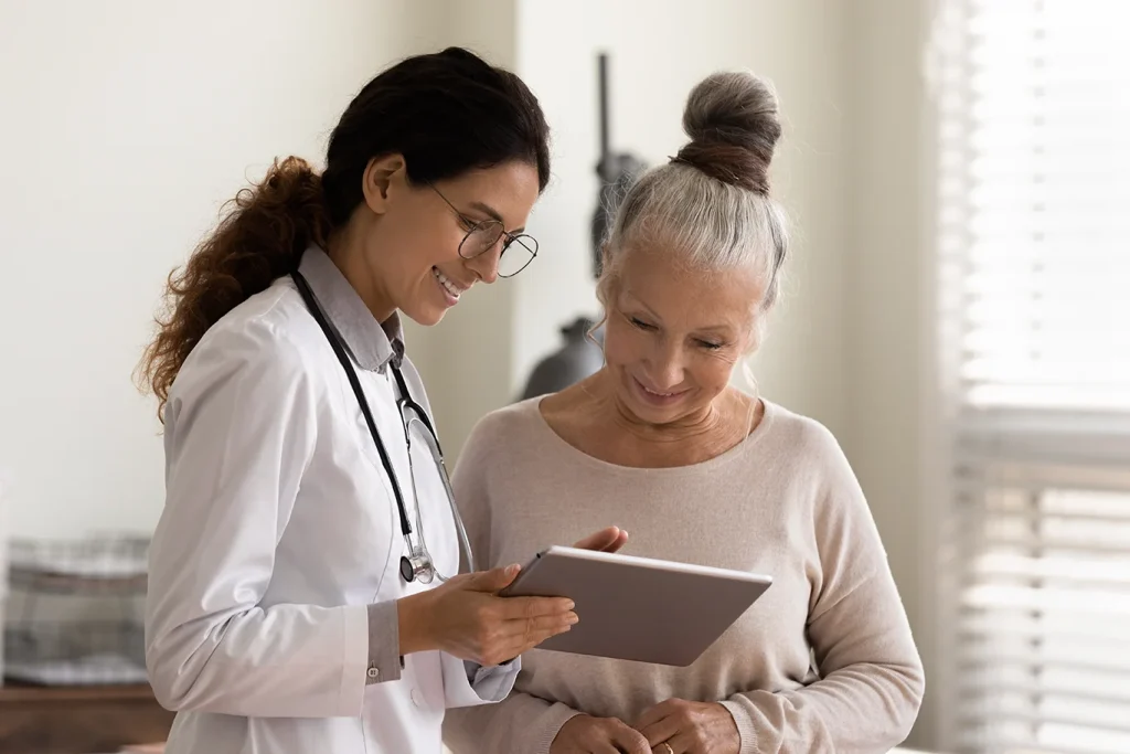 Happy female doctor and senior patient look at tablet screen discuss treatment or therapy