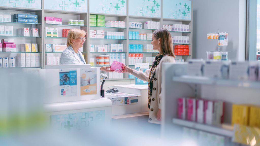 Pharmacy Drugstore Checkout Cashier Counter: Mature Female Pharmacist Passes Pink Box with Cure to a Female Customer, who is Buying Prescription Medicine, Vitamins, Beauty, Health Care Products.