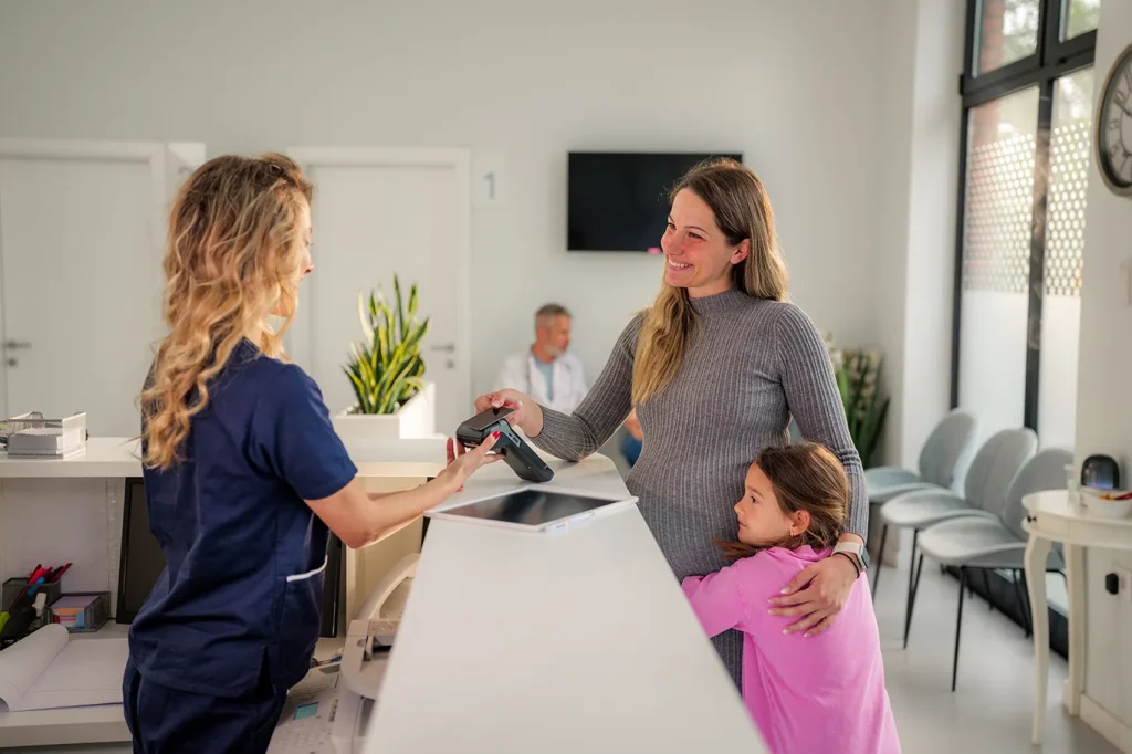 Pregnant woman paying with contactless payment at the clinic reception, while her daughter is hugging her